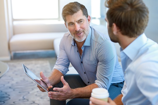 A mature business man sitting in a business lounge with a young man, offering mentoring advice to him while holding a digital tablet