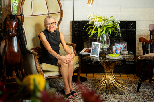 Margaret Dickson is seen sitting on a chair in her living room, next to a fireplace adorned with art.