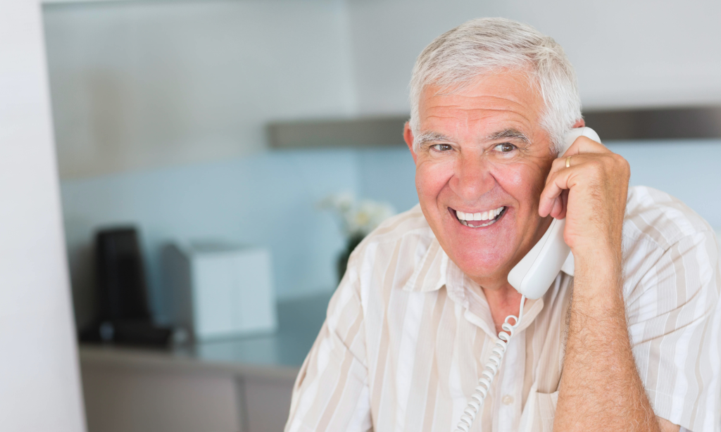 An adult man smiles as he talks on a white corded telephone.