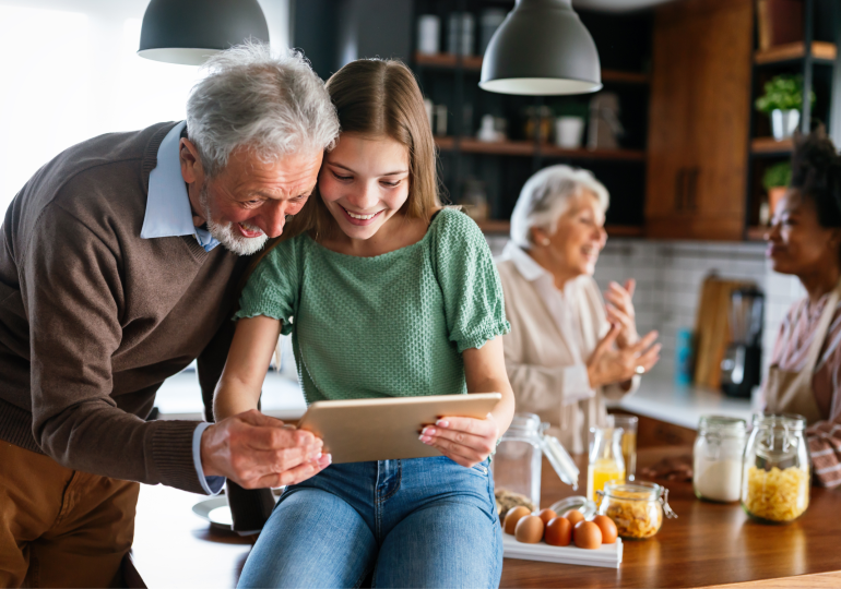 A man and a woman looking at a tablet together in a kitchen, with two women talking in the background.