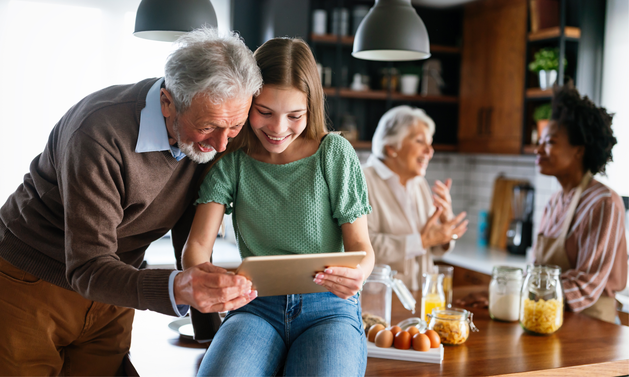 A man and a woman looking at a tablet together in a kitchen, with two women talking in the background.